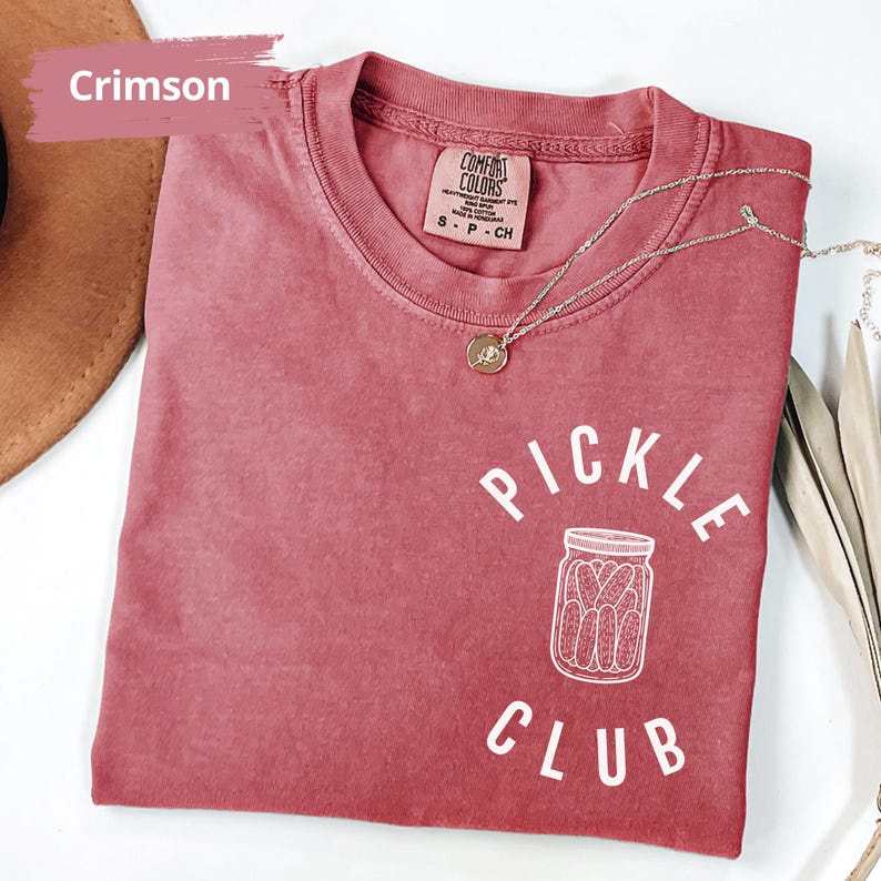 Peut inclure: Un t-shirt rouge cramoisi avec l'inscription "Pickle Club" en blanc. Le t-shirt pr&eacute;sente une illustration blanche d'un pot de cornichons.