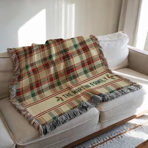 May include: A woven throw blanket featuring a red, green, and cream plaid pattern with fringed edges. The blanket includes a horizontal cream-colored band with the text THE MARTIN FAMILY in green, accented by small illustrations of holly berries.