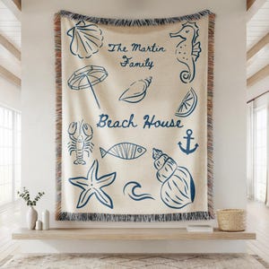 May include: A beige beach-themed throw blanket with blue illustrations of seashells, a seahorse, and other nautical elements. The text "The Martin Family Beach House" is also displayed on the blanket.
