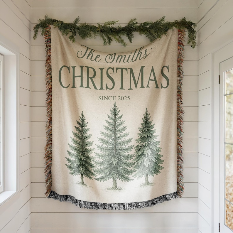 以下が含まれることがあります： 「The Smiths' CHRISTMAS SINCE 2025」の文字と3本の緑のクリスマスツリーが描かれたベージュ色のクリスマス用スローケット。フリンジ付きの縁があり、緑のガーランドで飾られた白い壁に掛けられています。