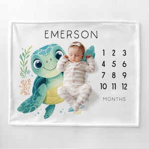 May include: White baby milestone blanket with a watercolor illustration of a smiling sea turtle and the name "EMERSON" in black. The blanket includes numbers 1-12 and the word "MONTHS" for tracking baby's growth.