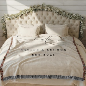 Op de afbeelding: Een cr&egrave;mekleurige sprei met zwarte tekst "Kaylee & Lennox EST 2025" ligt op een bed. De sprei heeft een franjerand en een decoratief ontwerp. Het hoofdbord is gecapitonneerd en er is een slinger overheen gedrapeerd.