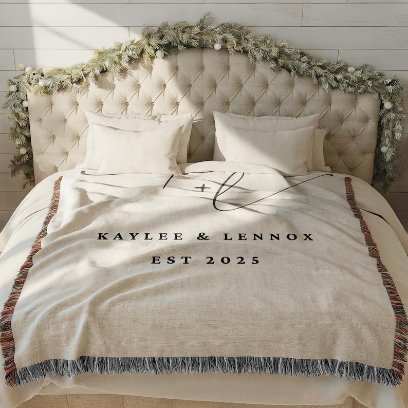 Op de afbeelding: Een cr&egrave;mekleurige sprei met zwarte tekst "Kaylee & Lennox EST 2025" ligt op een bed. De sprei heeft een franjerand en een decoratief ontwerp. Het hoofdbord is gecapitonneerd en er is een slinger overheen gedrapeerd.