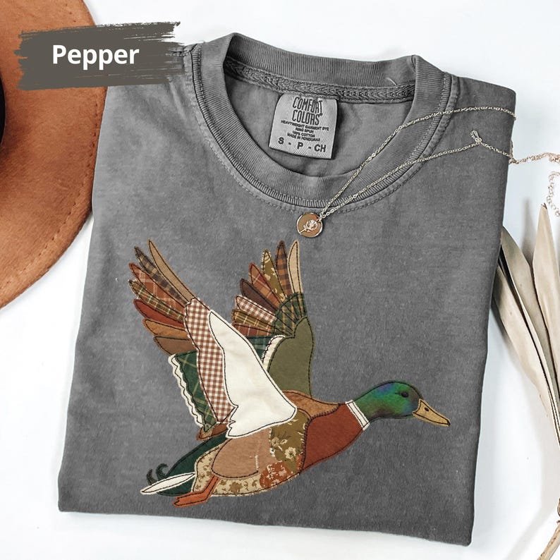 K&ouml;nnte beinhalten: Ein graues Comfort Colors T-Shirt mit einem Patchwork-Enten-Design im Flug. Die Ente hat braune, gr&uuml;ne und wei&szlig;e gemusterte Fl&uuml;gel und einen K&ouml;rper. Eine silberne Halskette mit einem kleinen Anh&auml;nger liegt auf dem Shirt. Das Wort "Pepper" befindet sich oben links.