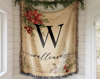 May include: A beige throw blanket with a large black letter "W" and the name "williams" in script. The blanket features a poinsettia and holly design with a fringed edge. A garland of greenery hangs above.