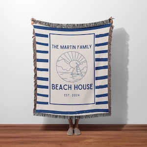 May include: A woven throw blanket with blue and white stripes. The blanket features a logo with a sailboat, sun, and clouds in a circle. The text "The Martin Family Beach House Est. 2024" is printed on the blanket.