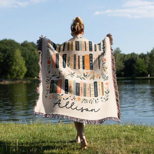 May include: A cream-colored woven throw blanket with a pattern of books and floral accents. The blanket has a fringe border and the name "Allison" is woven into the bottom center.
