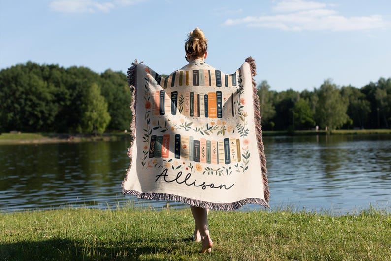 May include: A cream-colored woven throw blanket with a pattern of books and floral accents. The blanket has a fringe border and the name "Allison" is woven into the bottom center.