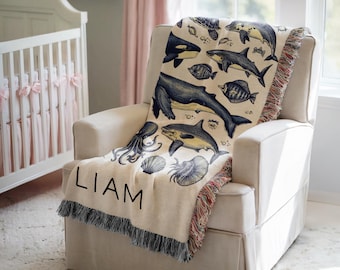 May include: A cream-colored woven throw blanket with a navy blue and white ocean animal design. The blanket has a fringe border and the name "LIAM" is printed in black on the front.