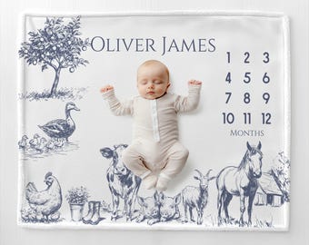 May include: A white baby milestone blanket with a navy blue farm animal illustration and the name "Oliver James". The blanket features a duck, cow, horse, and other farm animals. Numbers 1-12 are printed on the right side.