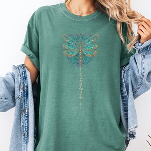 May include: Teal green t-shirt featuring a gold dragonfly mandala design with the word "wings" written vertically in gold script.