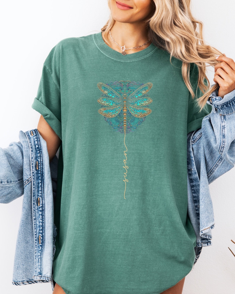 May include: Teal green t-shirt featuring a gold dragonfly mandala design with the word "wings" written vertically in gold script.