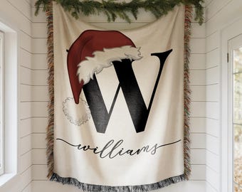 May include: A cream-colored woven throw blanket featuring a large black capital letter W topped with a red and white Santa hat. Below the letter, the name Williams is printed in a black cursive script. The blanket has fringed edges on all sides.