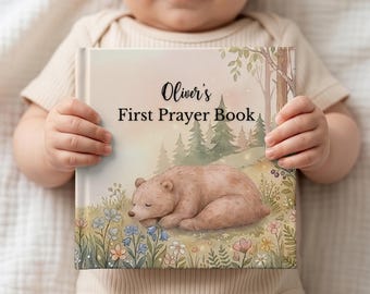 Puede incluir: Un libro infantil titulado "First Prayer Book de Oliver" sostenido por un bebé. La portada presenta una ilustración en acuarela de un oso pardo durmiendo en un campo de flores, con árboles al fondo. El libro tiene una paleta de colores pastel.