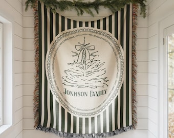 May include: A green and white striped throw blanket with a fringed edge. The blanket features a circular design with a Christmas tree and the text "JONHSON FAMILY". A garland of greenery is draped above the blanket.