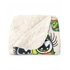 May include: A plush throw blanket with a cartoon pickle design. The pickle has large eyes, eyelashes, and a heart-shaped pupil. The pickle is green and has a red blush on its cheek. There is a jar of pickles in the background.