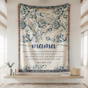 May include: A beige throw blanket with a blue floral pattern and fringe trim. The word "mama" is written in cursive, with a quote below. The quote reads "She is clothed with strength and love... Proverbs 31:25."