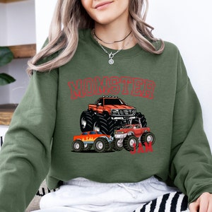 May include: A green sweatshirt with a red graphic of monster trucks. The graphic says "Monster Jam" and features three monster trucks, one of which is orange and the other two are red.