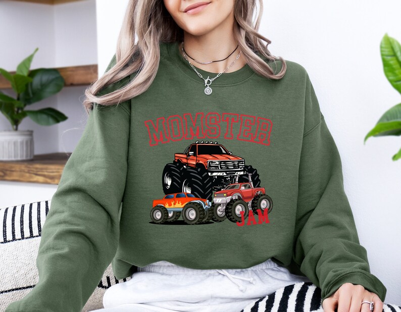 May include: A green sweatshirt with a red graphic of monster trucks. The graphic says "Monster Jam" and features three monster trucks, one of which is orange and the other two are red.