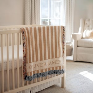 May include: A tan and white striped baby blanket with the name "OLIVER JAMES" woven into the fabric. The blanket is draped over the side of a white crib. The blanket has a fringed edge.