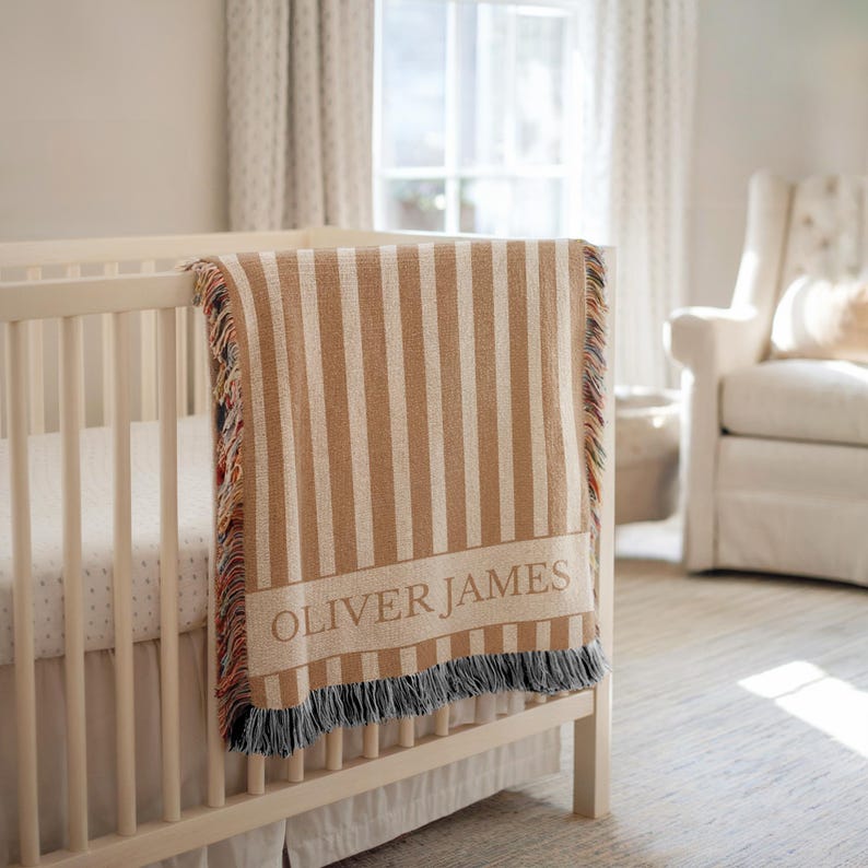 May include: A tan and white striped baby blanket with the name "OLIVER JAMES" woven into the fabric. The blanket is draped over the side of a white crib. The blanket has a fringed edge.