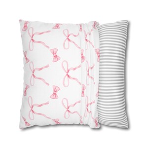 Puede incluir: Una almohada blanca con un patr&oacute;n de lazos rosas en un lado y un patr&oacute;n de rayas grises y blancas en el otro.