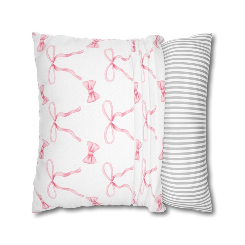 Puede incluir: Una almohada blanca con un patr&oacute;n de lazos rosas en un lado y un patr&oacute;n de rayas grises y blancas en el otro.