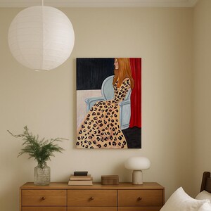 Puede incluir: Escena interior con una pintura de una mujer con un vestido con estampado de leopardo sentada en una silla azul claro. La obra de arte cuelga sobre una cómoda de madera con una lámpara, libros y un jarrón con vegetación. Una linterna de papel blanco cuelga del techo.