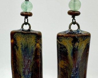 May include: A pair of handcrafted earrings with a unique design. Each earring features a rectangular, glazed ceramic pendant with a blend of brown, blue, and yellow hues. The earrings also include a green bead and a dark metal hook.