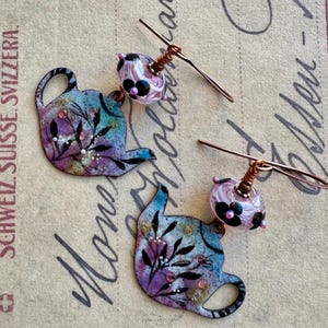 May include: Handmade earrings featuring a teapot design. The earrings have a blue, purple, and gold color scheme with floral accents. Each earring has a glass bead with black floral details and copper wire.