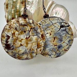 May include: A pair of round earrings with a cracked, mosaic-like pattern in shades of brown, beige, and gray. The earrings are suspended from dark metal hooks and are displayed against a seashell background.