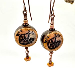 May include: Copper-colored dangle earrings featuring a round, painted bead with a black sloth design. The earrings have copper wire accents and small orange beads, hanging from a copper hook. The earrings are hanging from a brown branch.