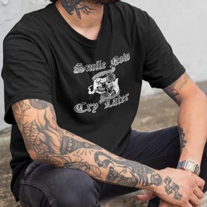 Może przedstawiać: Czarny T-shirt z białym nadrukiem graficznym, na którym widnieje napis "Smile Now Cry Later" z motywem czaszki i skrzyżowanych kości.