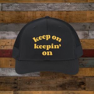 May include: A black trucker hat with a yellow embroidered message that reads "keep on keepin' on".