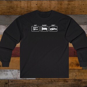 May include: Black long-sleeve t-shirt with white text that reads "Eat Sleep Drift" and three icons: a fork and knife, a bed, and a car drifting.
