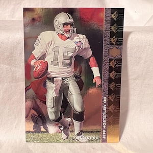 Puede incluir: Una tarjeta de fútbol coleccionable con Jeff Hostetler, mariscal de campo de los Raiders. La tarjeta muestra a Hostetler con su uniforme de los Raiders, sosteniendo un balón de fútbol. La tarjeta tiene un borde dorado y verde con el texto "SP" y "JEFF HOSTETLER QB".