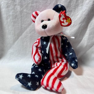 May include: A plush teddy bear with a patriotic design. The bear's body is split, with one side featuring red and white stripes and the other with a blue background and white stars. The bear has a pink face, ears, and a black nose.