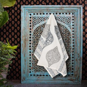May include: A white and gray patterned tea towel hangs on a distressed turquoise wooden frame with intricate cut-out designs. The towel features large leaf-like motifs and geometric borders, set against a black and gold patterned backdrop.