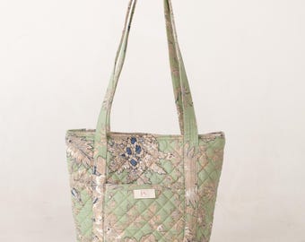 Puede incluir: Un bolso tote acolchado con un estampado floral en tonos verdes, beige y azul. El bolso tiene dos asas y un pequeño bolsillo delantero. El bolso mide aproximadamente 36 cm de alto y 30 cm de ancho.
