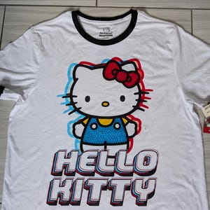 May include: White t-shirt with a graphic of Hello Kitty in a blue and white outfit with a red bow. The text "Hello Kitty" is in a 3D style with red, blue, and white accents.