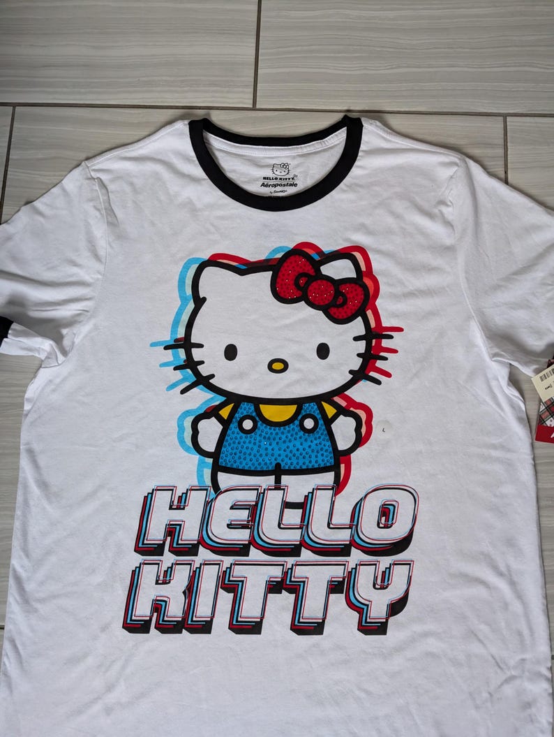 May include: White t-shirt with a graphic of Hello Kitty in a blue and white outfit with a red bow. The text "Hello Kitty" is in a 3D style with red, blue, and white accents.