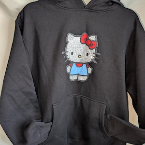 May include: Black hoodie featuring a Hello Kitty graphic. The graphic shows Hello Kitty with a red bow, blue overalls, and a white body. The sweatshirt has a front pocket and a drawstring hood.