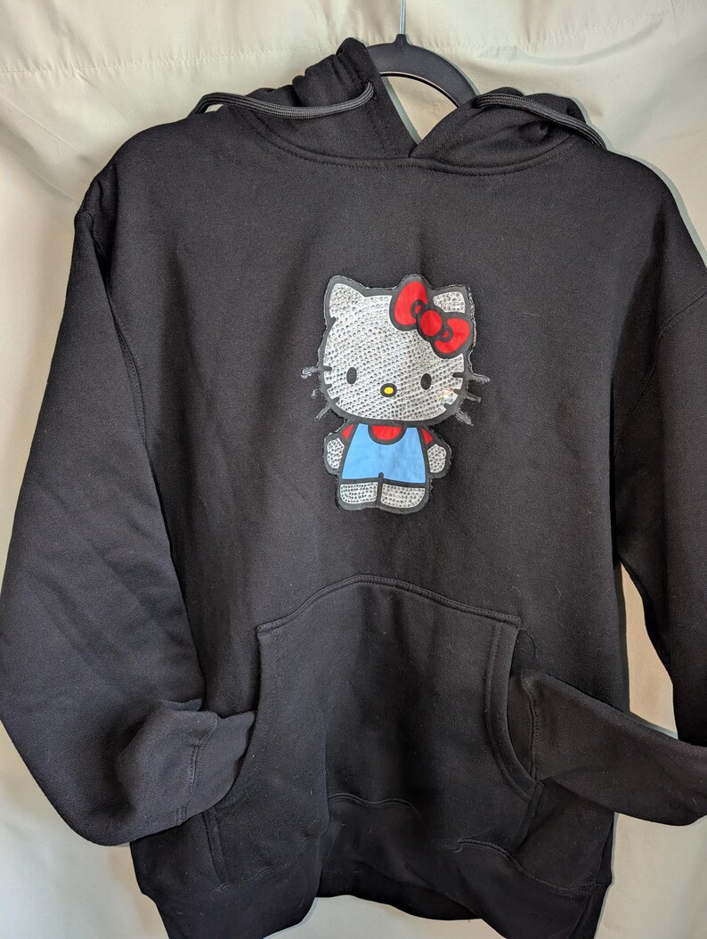 May include: Black hoodie featuring a Hello Kitty graphic. The graphic shows Hello Kitty with a red bow, blue overalls, and a white body. The sweatshirt has a front pocket and a drawstring hood.