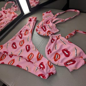 May include: Pink bikini with a red lip print. The bikini top has a halter style with adjustable straps. The bikini bottom is a high-cut style.