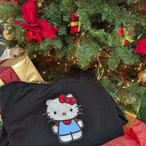 May include: A black sweatshirt with a sequined Hello Kitty patch, featuring a red bow and blue overalls. The sweatshirt is placed in front of a decorated Christmas tree with baubles and lights, creating a festive holiday scene.