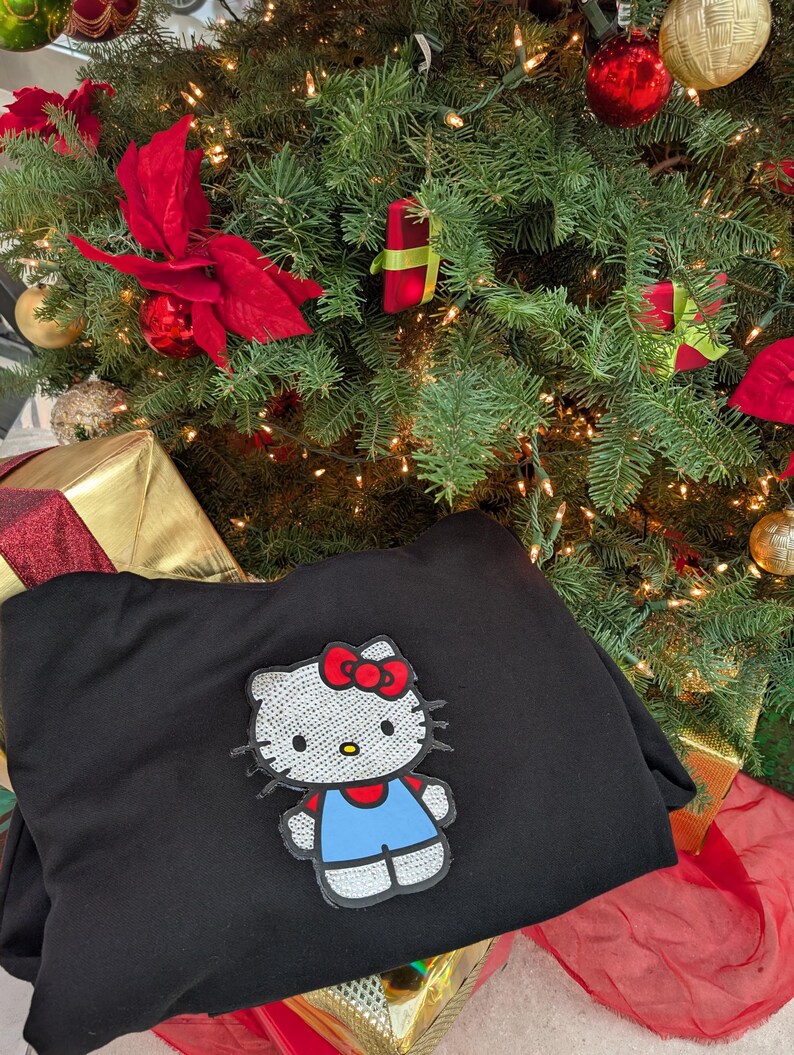 May include: A black sweatshirt with a sequined Hello Kitty patch, featuring a red bow and blue overalls. The sweatshirt is placed in front of a decorated Christmas tree with baubles and lights, creating a festive holiday scene.