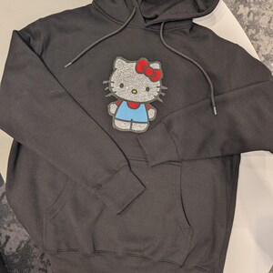 May include: Black hooded sweatshirt with a front pocket and drawstring. The sweatshirt features a sparkly Hello Kitty graphic with a red bow, blue overalls, and a white face.