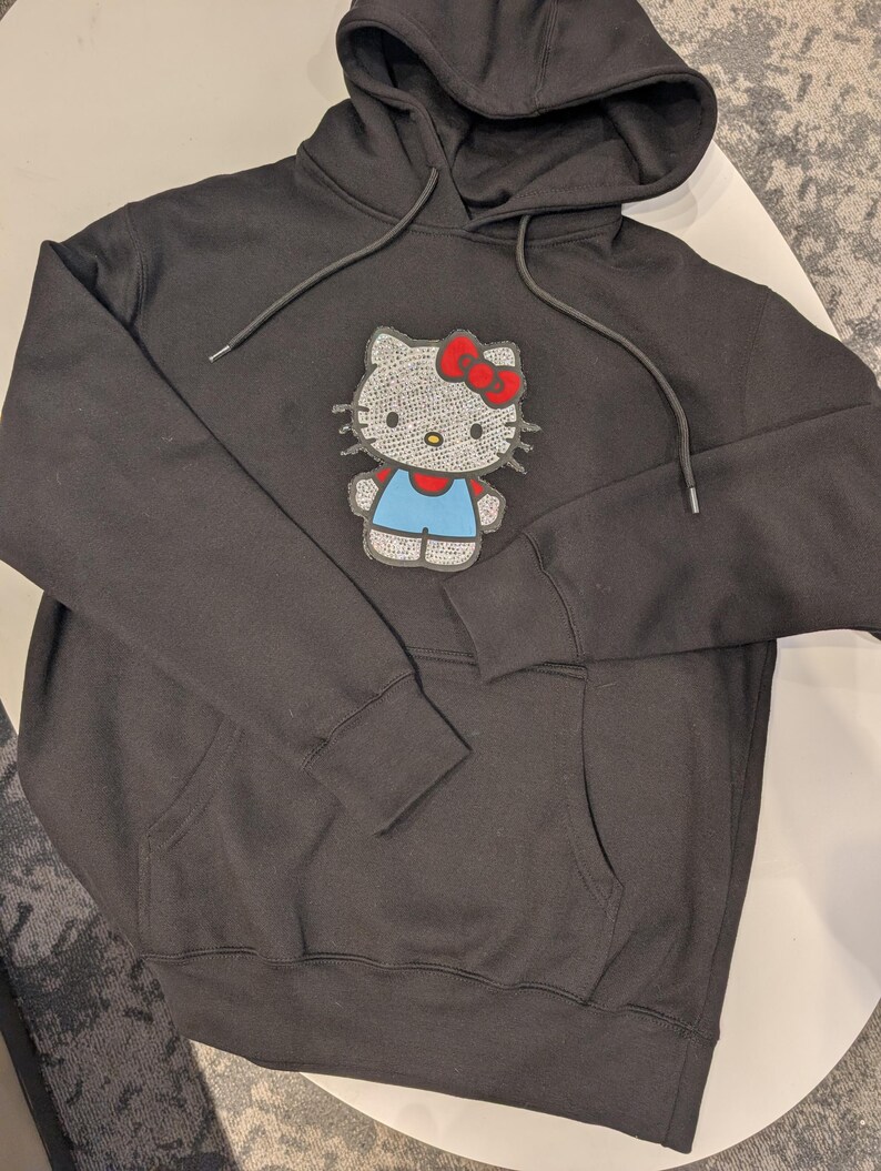 May include: Black hooded sweatshirt with a front pocket and drawstring. The sweatshirt features a sparkly Hello Kitty graphic with a red bow, blue overalls, and a white face.