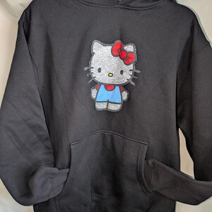 May include: Black hooded sweatshirt with a Hello Kitty graphic. The graphic shows Hello Kitty with a red bow, blue overalls, and a silver sequined body. The sweatshirt has a front pocket and a drawstring hood.
