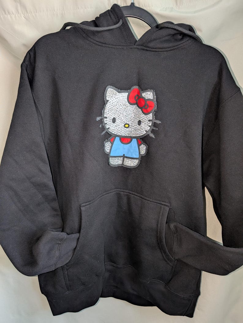 May include: Black hooded sweatshirt with a Hello Kitty graphic. The graphic shows Hello Kitty with a red bow, blue overalls, and a silver sequined body. The sweatshirt has a front pocket and a drawstring hood.